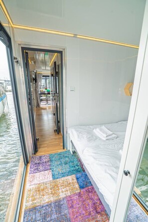 A narrow corridor with a bed on the right, a colorful rug, and a view of the water outside.