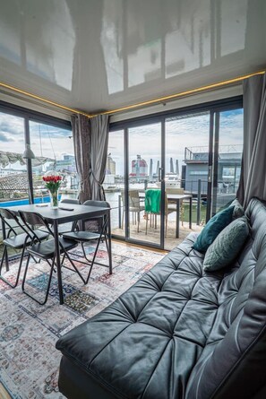 A modern living room with a leather sofa, a dining table with chairs, and a large window offering a city view.