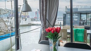 Et værelse med et bord, en vase med blomster, en lampe og udsigt til en havnefront med bygninger.