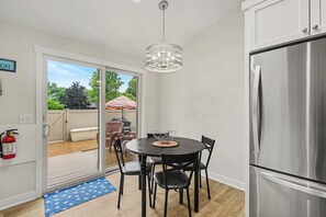 Eine Küche mit einem Edelstahlkühlschrank, einem Esstisch mit Stühlen, einer Pendelleuchte und Blick ins Freie durch eine Schiebetür.