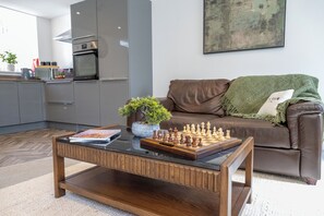 A modern kitchen with a built-in oven, a dining area with a chessboard, and a brown leather sofa with a green throw.