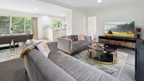Ein geräumiges Wohnzimmer mit grauem Sofa, Couchtisch und Fernseher. Im Hintergrund ist ein Balkon mit Essbereich und einer Küche zu sehen.