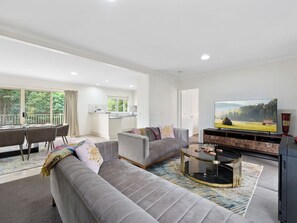 Ein geräumiges Wohnzimmer mit grauem Sofa, Couchtisch und Fernseher. Im Hintergrund ist ein Balkon mit Essbereich und einer Küche zu sehen.