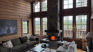 A cozy cabin interior with a stone fireplace, wooden walls, and large windows offering a view of the outdoors. - Luxury mountain lodge with mountain views and sauna (Idre)