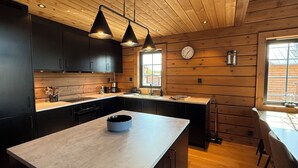 Een moderne keuken met houten wanden, zwarte kastjes, een centraal eiland, hanglampen en een klok aan de muur.
