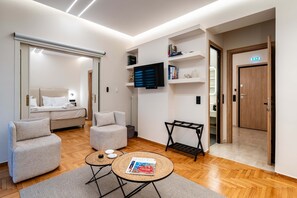 Quarto de hotel moderno com cama, dois sofás, uma mesinha redonda com uma revista, TV na parede e porta de madeira.
