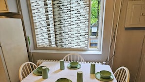 Una mesa de comedor con mantel blanco, platos verdes y cubiertos a juego, frente a una ventana con persiana estampada.