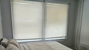 Un dormitorio con una cama, dos ventanas con persianas, un radiador y un enchufe en la pared.