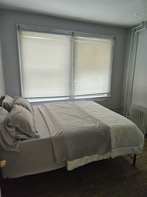 Une chambre à coucher comprenant un lit, deux fenêtres équipées de stores, un radiateur et une prise électrique fixée au mur.