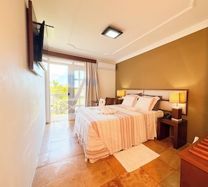 Quarto com cama, luminárias de cabeceira, mesa de apoio, janela com cortinas e vista para o verde lá fora.