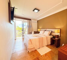 Quarto com cama, luminárias de cabeceira, mesa de apoio, janela com cortinas e vista para o verde lá fora.
