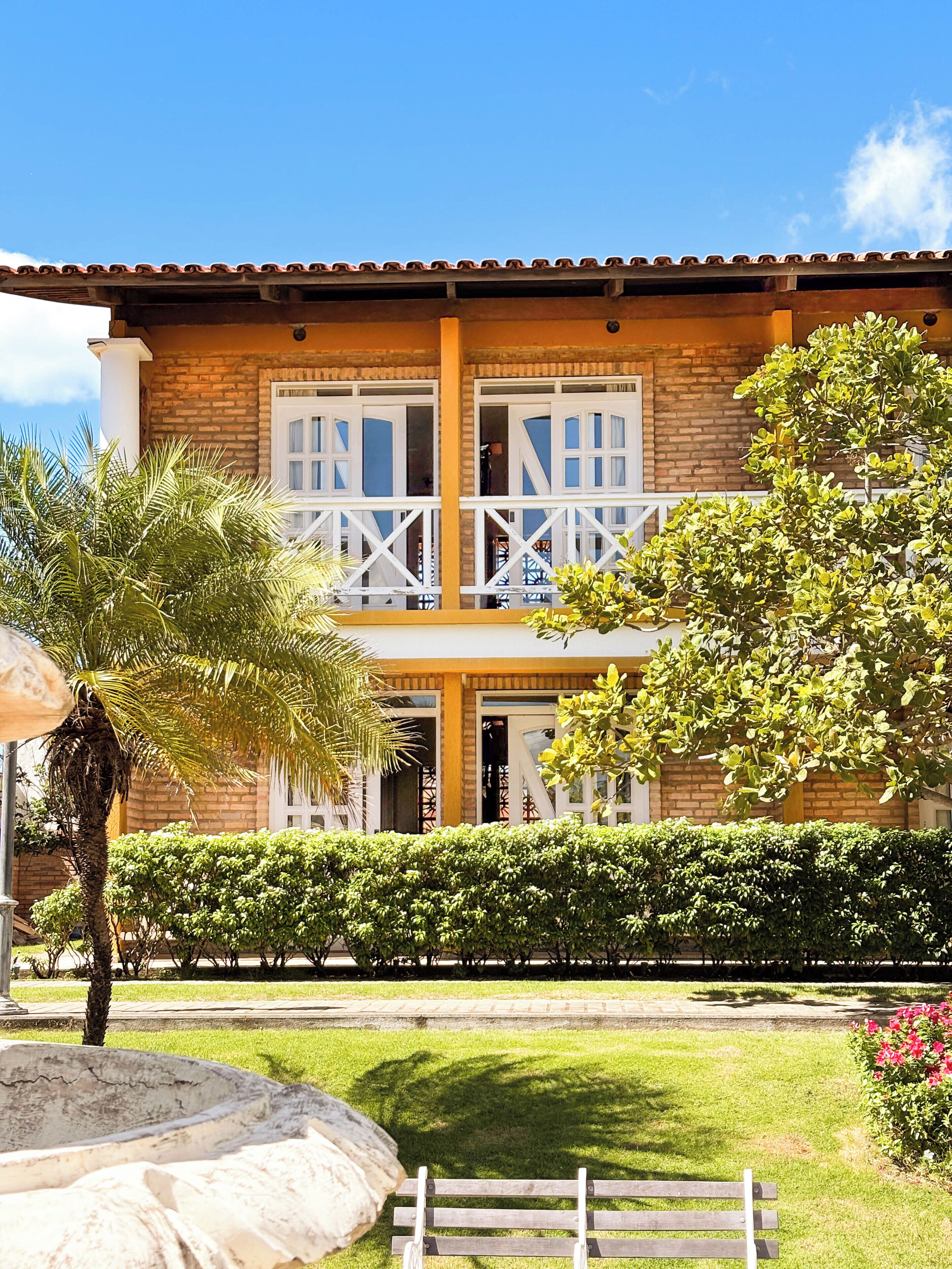 Um edifício de dois andares com fachada de tijolos, varanda com grades brancas e um jardim bem cuidado com um banco e uma palmeira.