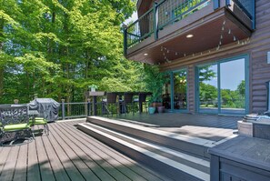 Eine Holzterrasse mit Gartenmöbeln, einem überdachten Grill und einem großen Fenster mit Blick auf einen bewaldeten Bereich.