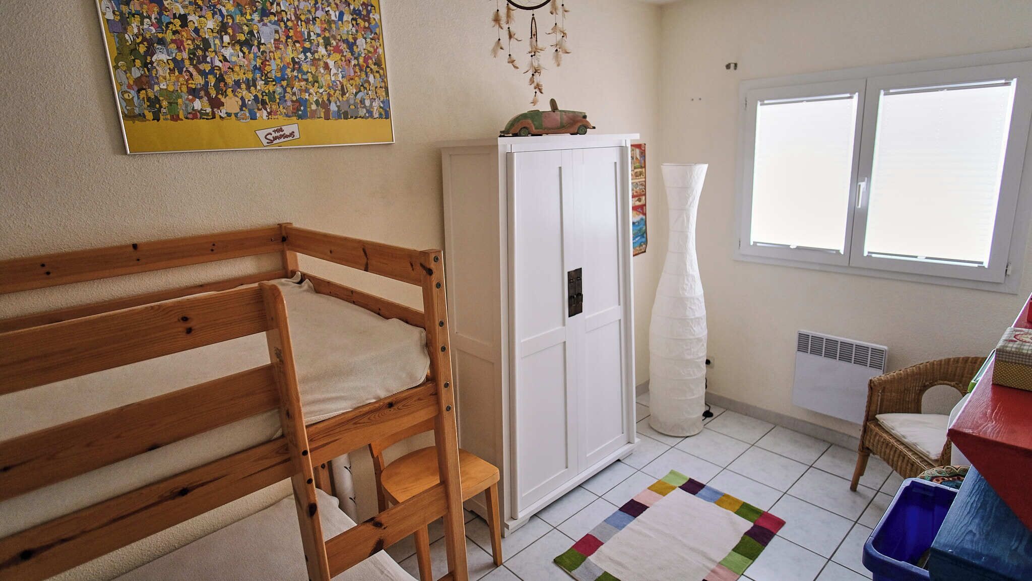 Une chambre avec un lit superposé en bois, une armoire blanche, un tapis coloré, une chaise en osier et une fenêtre avec des stores.