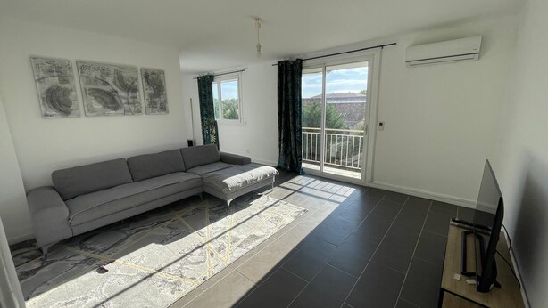 A living room with a grey sofa, a flat-screen TV on a wooden stand, a large window with a view, and a balcony. - Spacious apartment near downtown (Avignon)