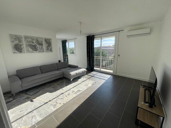 Ein Wohnzimmer mit grauem Sofa, einem Flachbildfernseher auf einem Holzboden, einem großen Fenster mit Aussicht und einem Balkon.