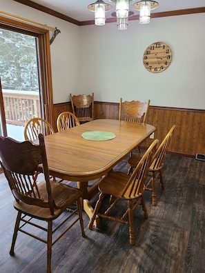 Une salle à manger avec une table et des chaises en bois, une horloge au mur et une fenêtre donnant sur des arbres.