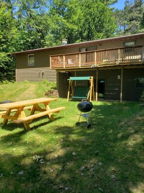 Une terrasse en bois avec une table de pique-nique et un barbecue, une maison avec un balcon et une cour gazonnée.