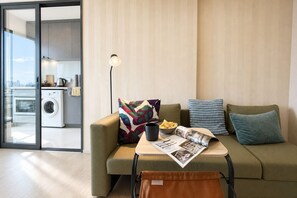 Ein modernes Wohnzimmer mit einer Couch, einem Couchtisch mit Zeitschriften, einer Lampe und einer Waschmaschine, die durch eine Glastür sichtbar ist.