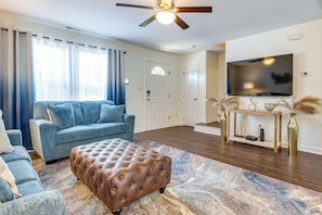 A living room with a blue sofa, a brown ottoman, a wooden console table with decorative items, and a mounted TV. - Great for Families! Raleigh Townhome 5 Mi to Dtwn (Raleigh)