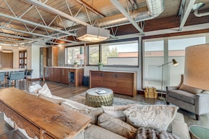 A spacious living area with a wooden bench, sofa, and armchair, a kitchen in the background, and large windows offering views. - Grand Haven Condo w/ Channel View & Dtwn Location (Grand Haven)
