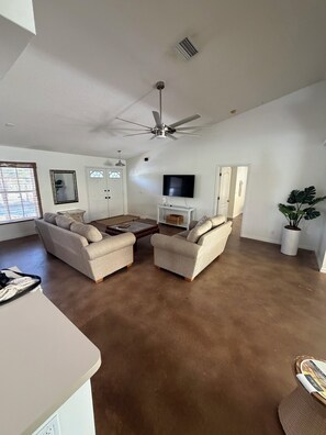 Ein geräumiges Wohnzimmer mit zwei beigefarbenen Sofas, einem Couchtisch, einem Deckenventilator, einem Fernseher und einer Topfpflanze.