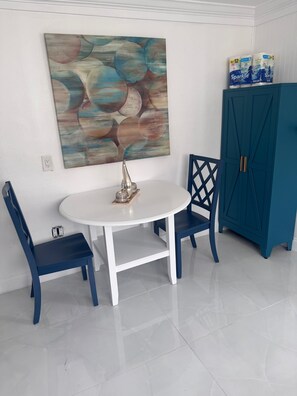 Ein Essbereich mit einem weißen Tisch, zwei blauen Stühlen, einem dekorativen Metallobjekt, einem blauen Schrank und einem großen, abstrakten Gemälde an der Wand.