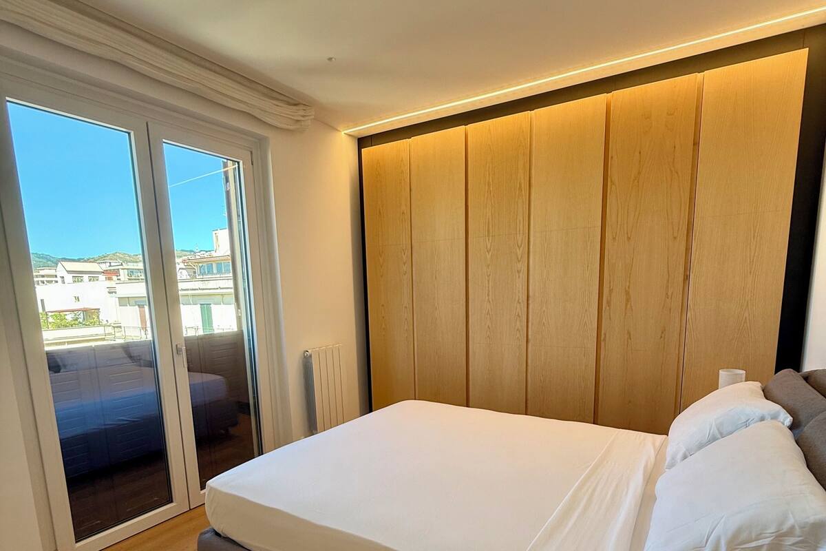 Une chambre dotée d’un grand lit, d’une tête de lit en bois, d’une table de chevet avec une lampe et d’une porte-fenêtre coulissante donnant sur un espace extérieur.