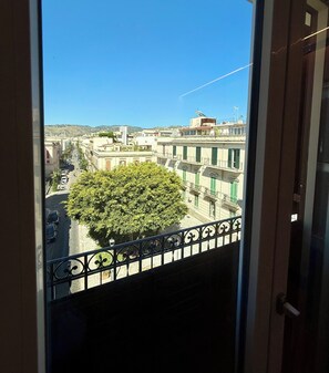 Un balcon doté d’une balustrade en fer forgé, donnant sur une rue bordée d’immeubles et d’un arbre.