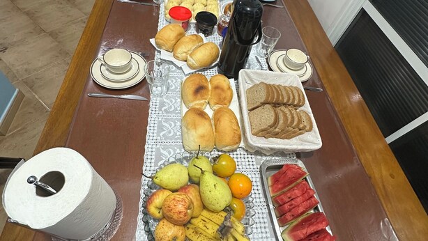 Una larga mesa de comedor con diversos alimentos y bebidas, una cafetera y un rollo de papel higiénico.
