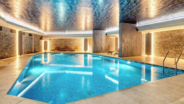 Ein überdachter Swimmingpool mit glasklar blauem Wasser, umgeben von Wänden mit steiniger Struktur und einer Decke mit Wolkenmuster.