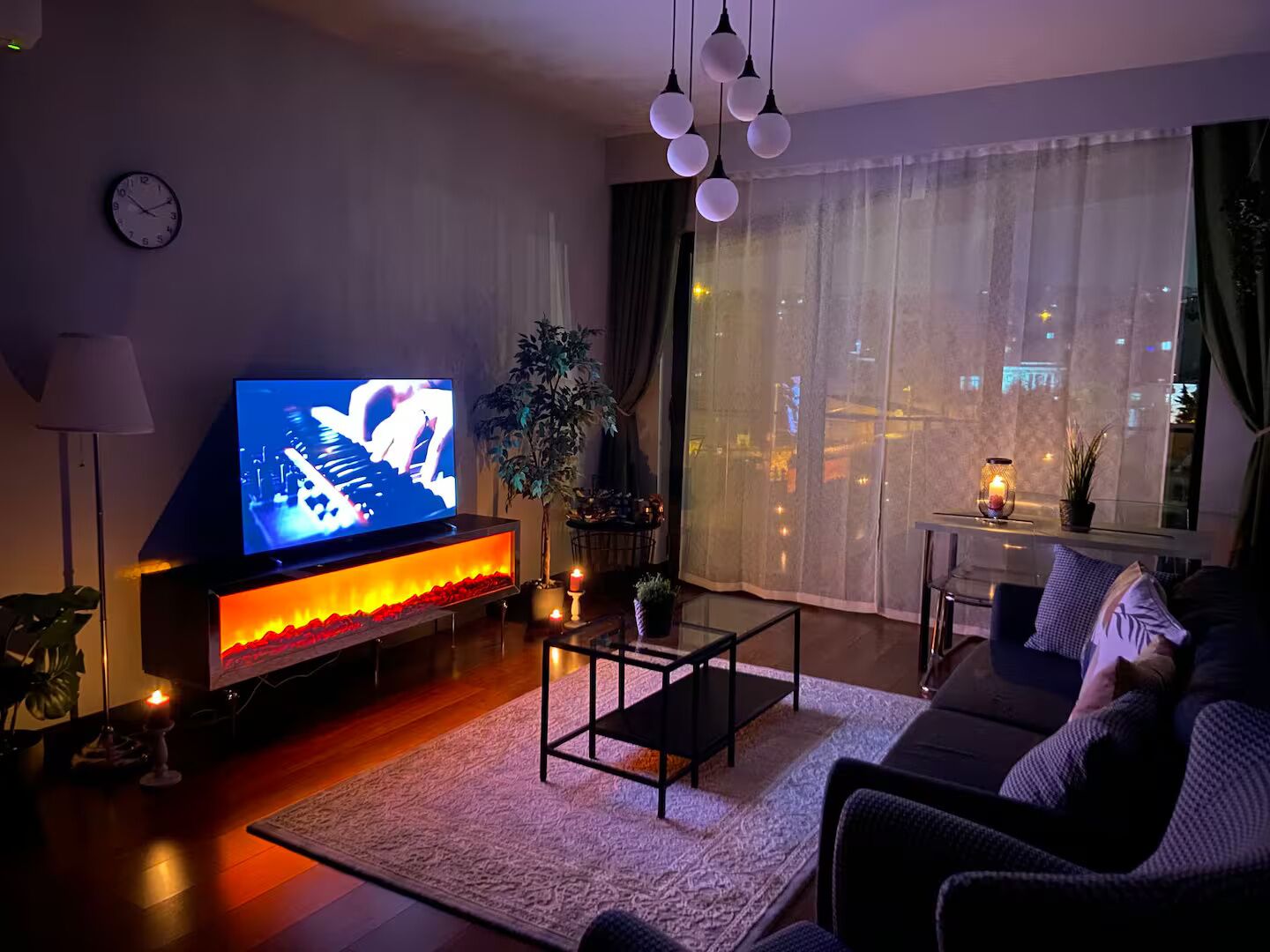 A modern living room with a flat-screen TV, a fireplace, a sofa, and a view of the city at night.