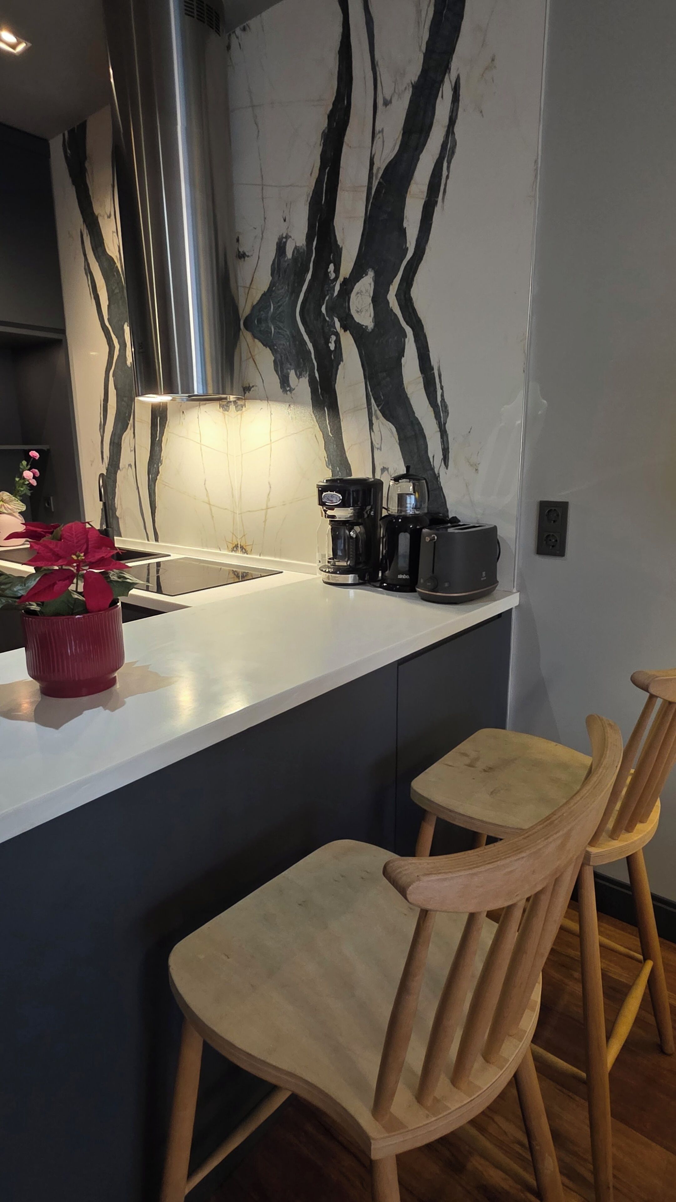 A modern kitchen with a marble backsplash, a stainless steel range hood, a coffee maker, and wooden chairs.