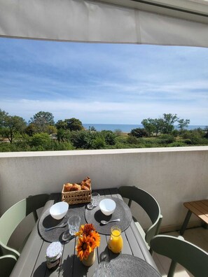 Une table dressée pour le petit-déjeuner, avec vue sur la mer, un panier de pain, un vase de fleurs et un verre de jus d’orange.