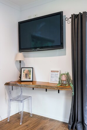 Een kamer met een wandtelevisie, bureau met lamp, stoel, een lijst met afbeelding, menukaart en een potplant.
