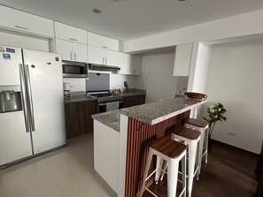 A modern kitchen with white cabinets, stainless steel appliances, a granite countertop, and wooden bar stools. - Apartment with a view of the Magdalena del Mar, Lima (Lima)