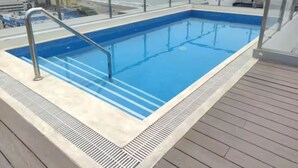 Una piscina rectangular con agua cristalina y azul, rodeada por una terraza de madera y una barandilla de vidrio.