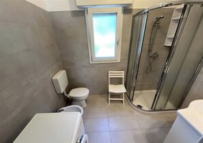 A bathroom with a toilet, a shower enclosure, a small window, and a white sink. - Quiet Apartment Near the sea (Lignano Sabbiadoro)