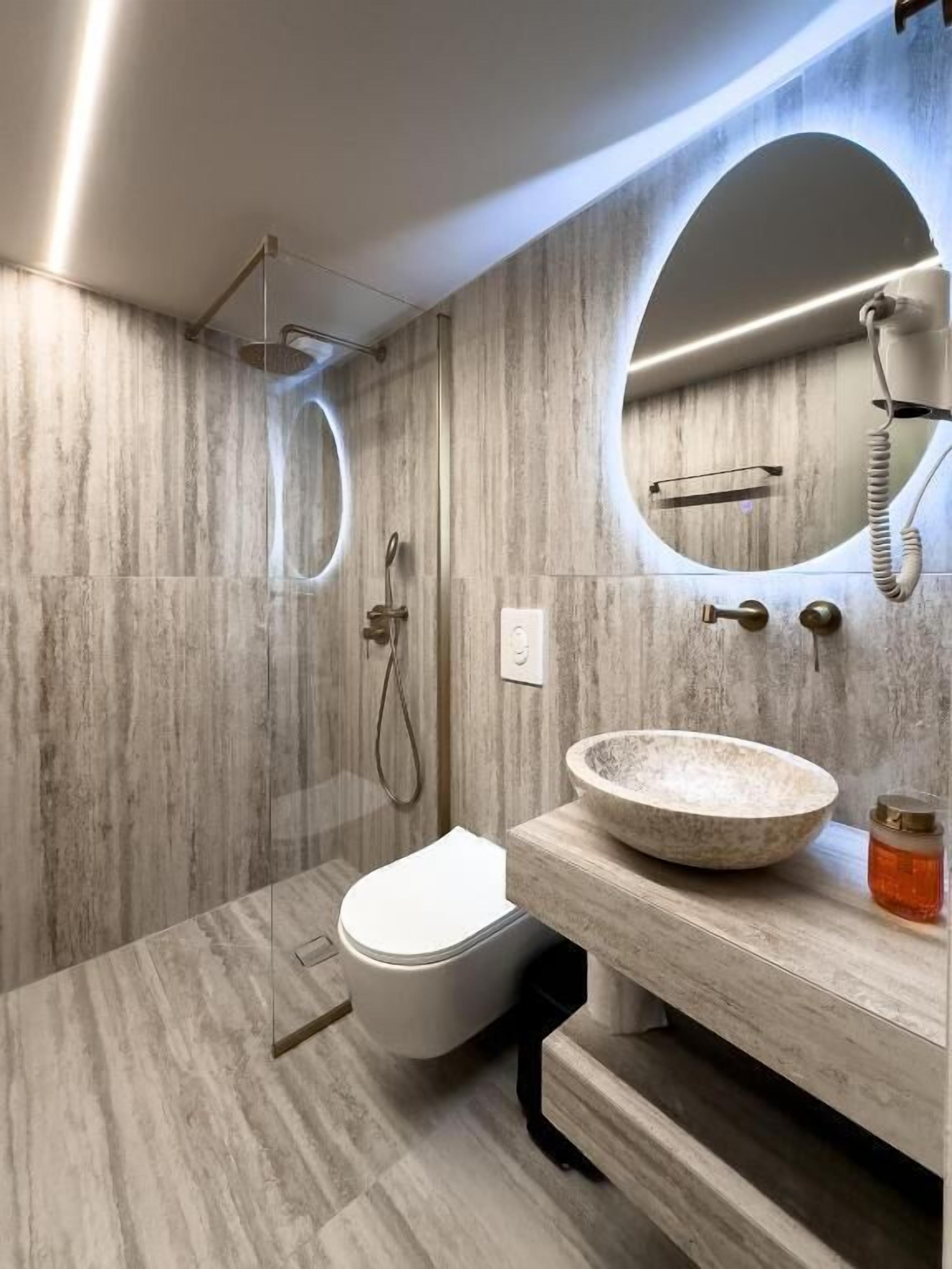Et moderne badeværelse med marmorgulv, et hvidt toilet, en rund vask med stentop og et spejl med telefon.