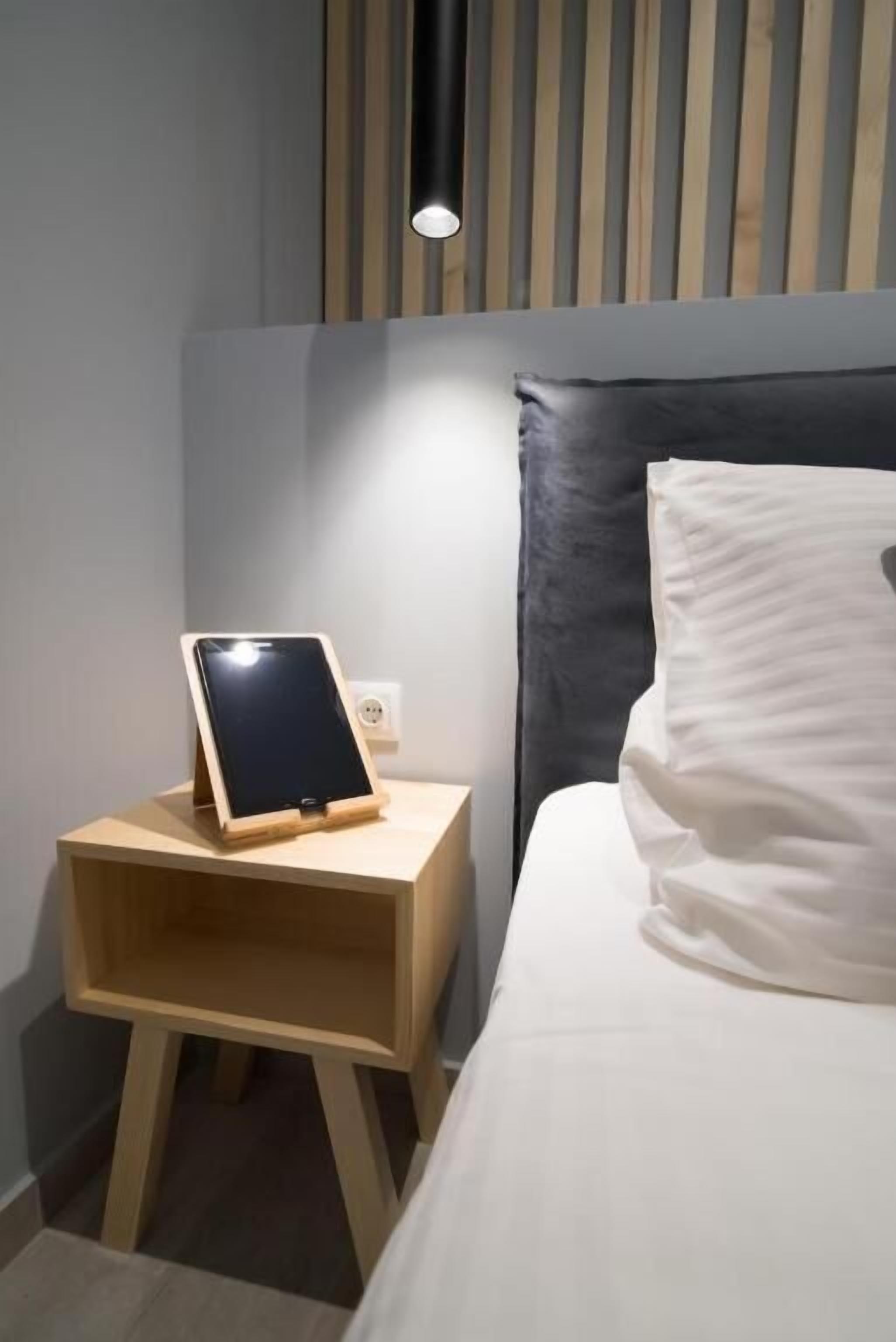 Una cama bien hecha con un cojín blanco y una mesita de noche de madera con una tableta.