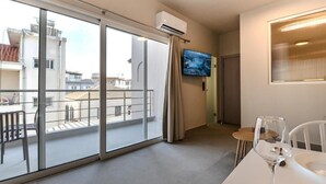 Una habitación de hotel moderna con balcón, televisor de pantalla plana y zona de comedor con mesa y sillas de vidrio.