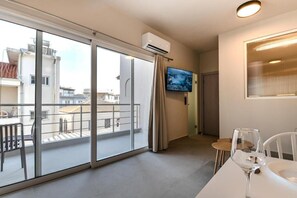 Una habitación de hotel moderna con balcón, televisor de pantalla plana y zona de comedor con mesa y sillas de vidrio.