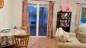 A living room with a sofa, a dining table with chairs, a bookshelf, and a sliding glass door leading to an outdoor area.