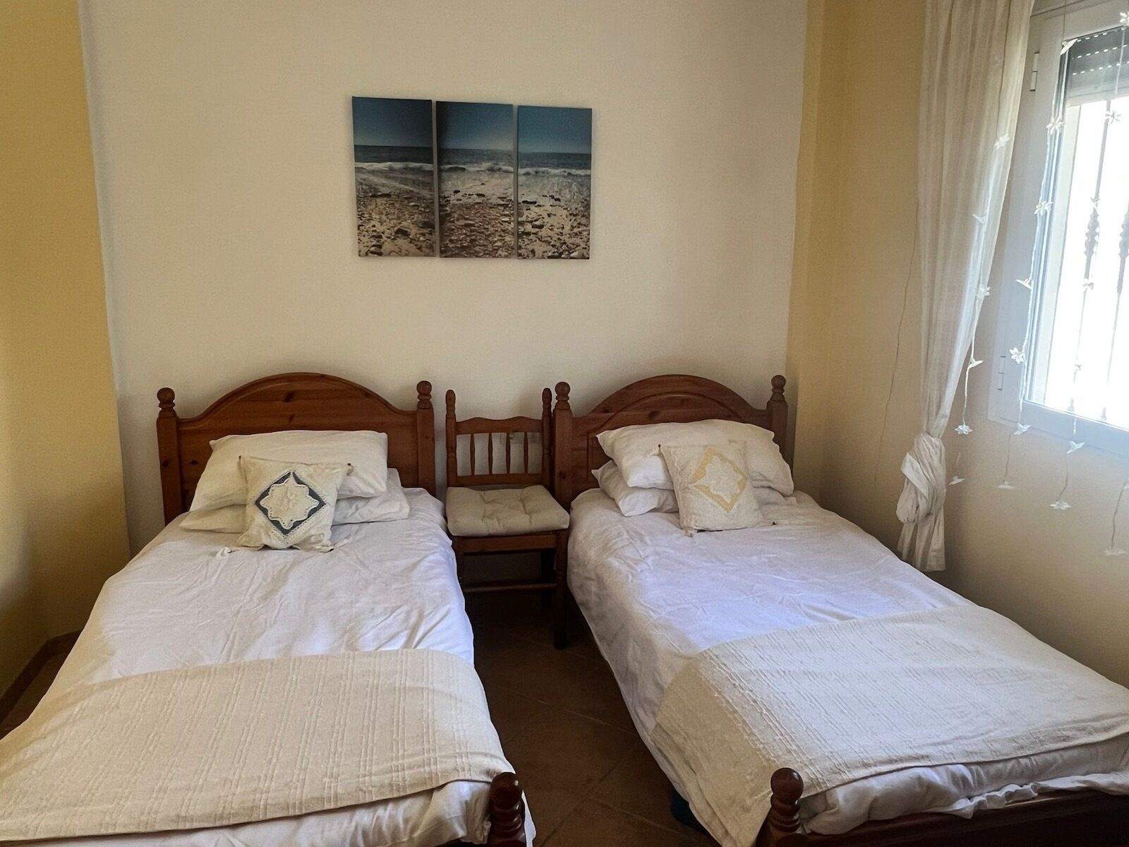 A bedroom with two wooden beds, white bedding, a wall-mounted artwork of a beach, and a window with curtains.