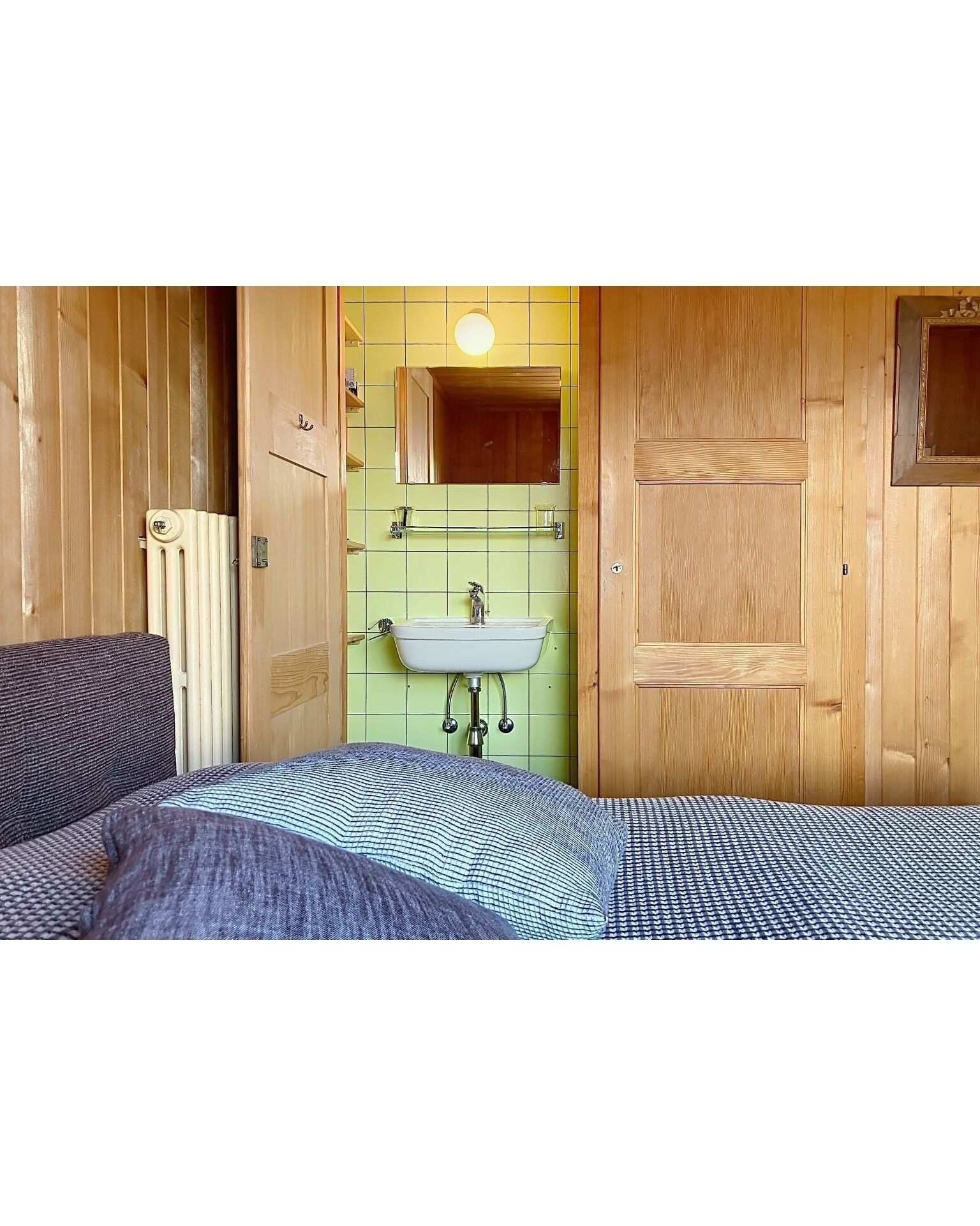Ein Zimmer mit einem Bett, einer Holztür, einer grünen Fliesenwand, einem weißen Waschbecken und einem Spiegel.