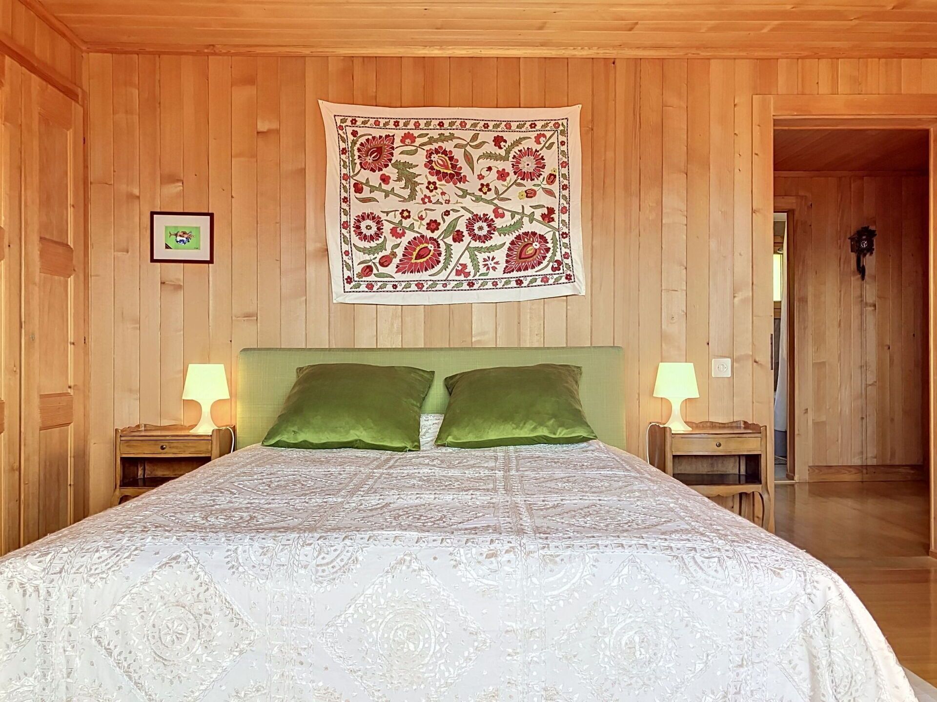 A wooden-paneled room with a bed, two nightstands, a tapestry, and a framed picture.