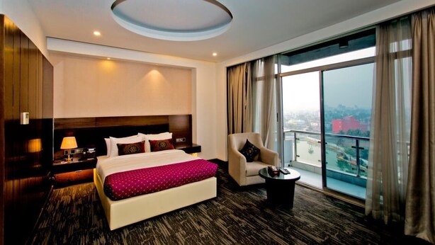 Ein Hotelzimmer mit einem großen Bett, einem Sessel, einem kleinen Tisch, einem Nachttisch und Blick auf die Stadt durch große Fenster.