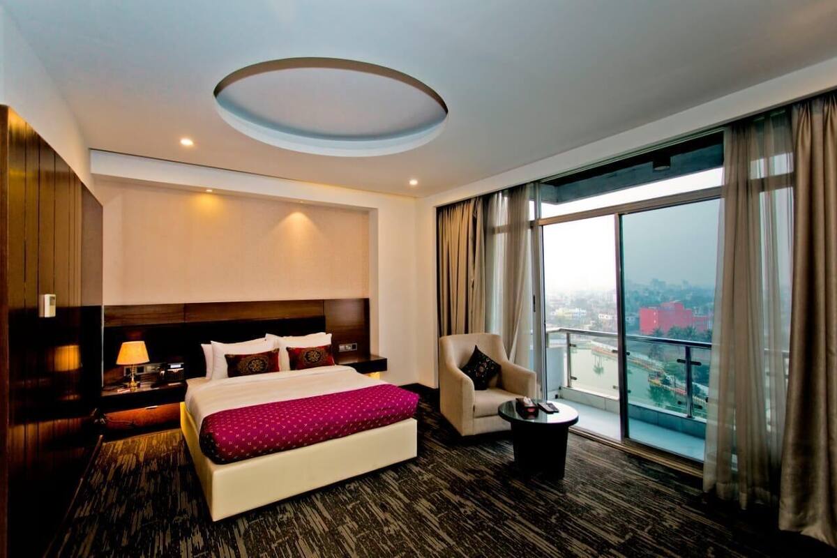 Quarto de hotel com uma cama grande, uma cadeira, uma mesinha, um criado-mudo e vista da cidade através de grandes janelas.