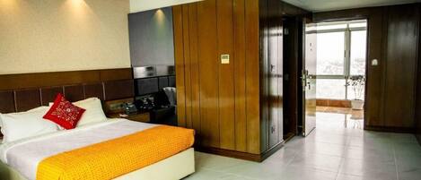 A hotel room with a bed, a wooden wardrobe, a desk with a chair, and a view of the outside.