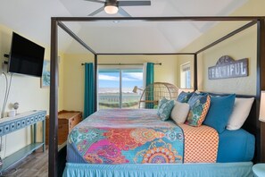 Un dormitorio con cama, televisor, ventilador de techo, ventana con cortinas y vistas al paisaje.